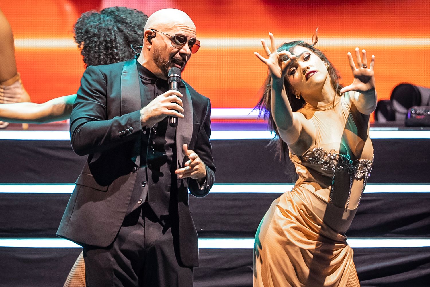 Bald male performer singing on stage with a female dancer in a gold outfit under dramatic red lighting.