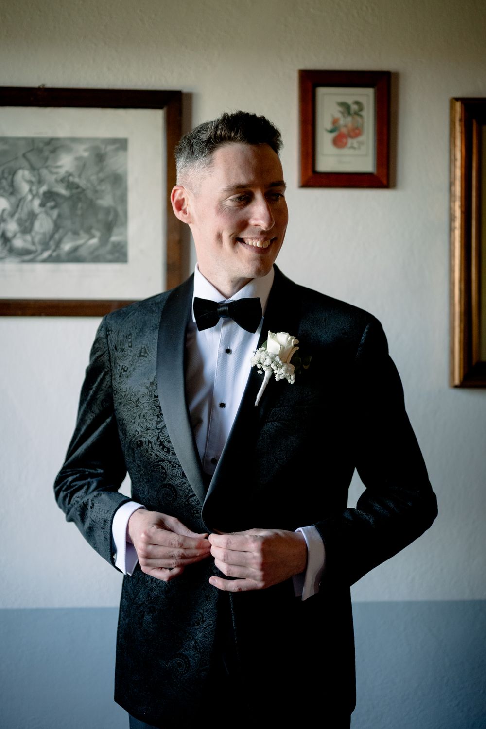 Smiling groom in black tuxedo with bow tie and white boutonniere buttoning jacket before wedding ceremony.