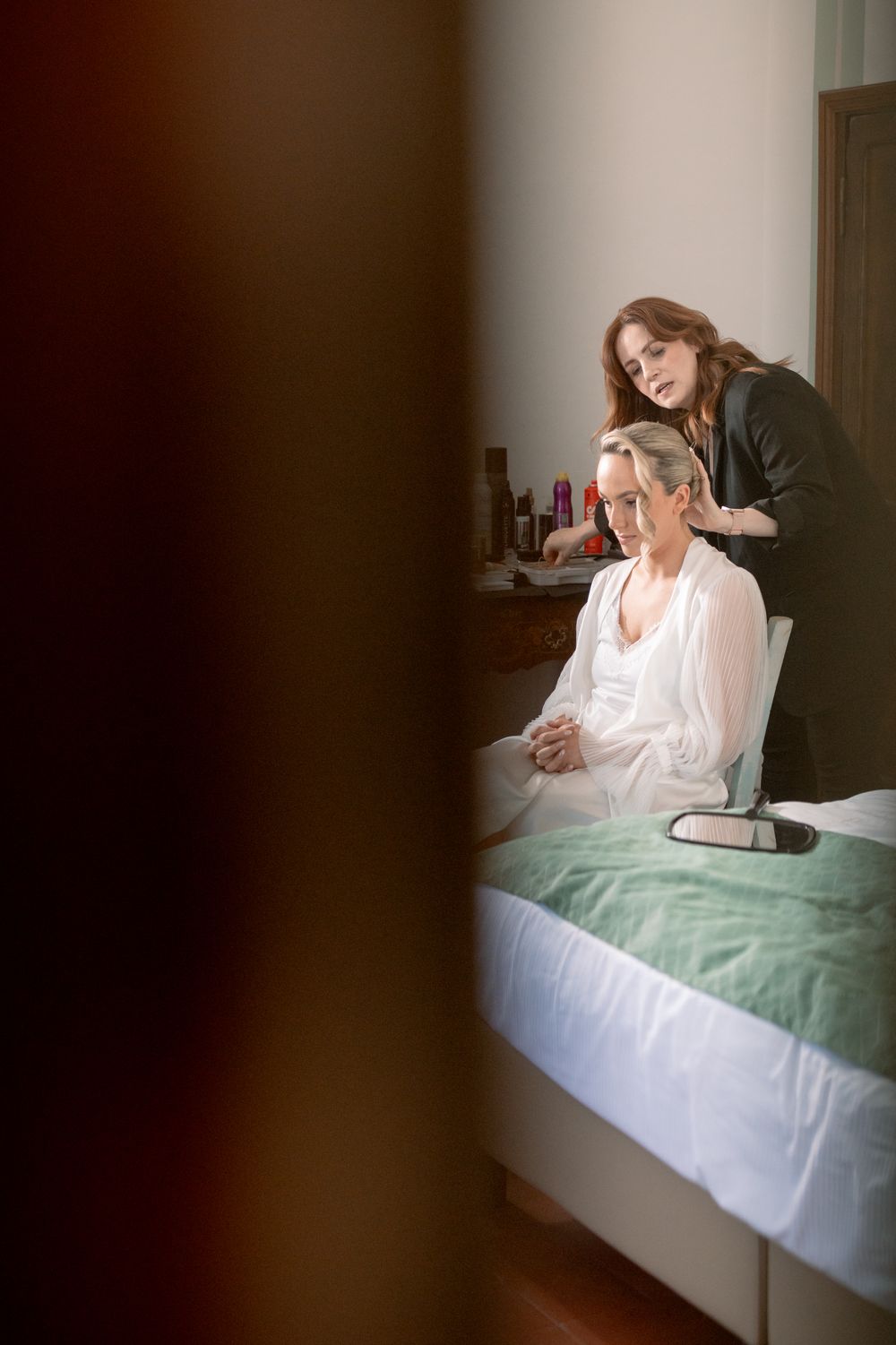 Hairstylist preparing bride's hair as she sits on bed in white robe during wedding morning preparations.