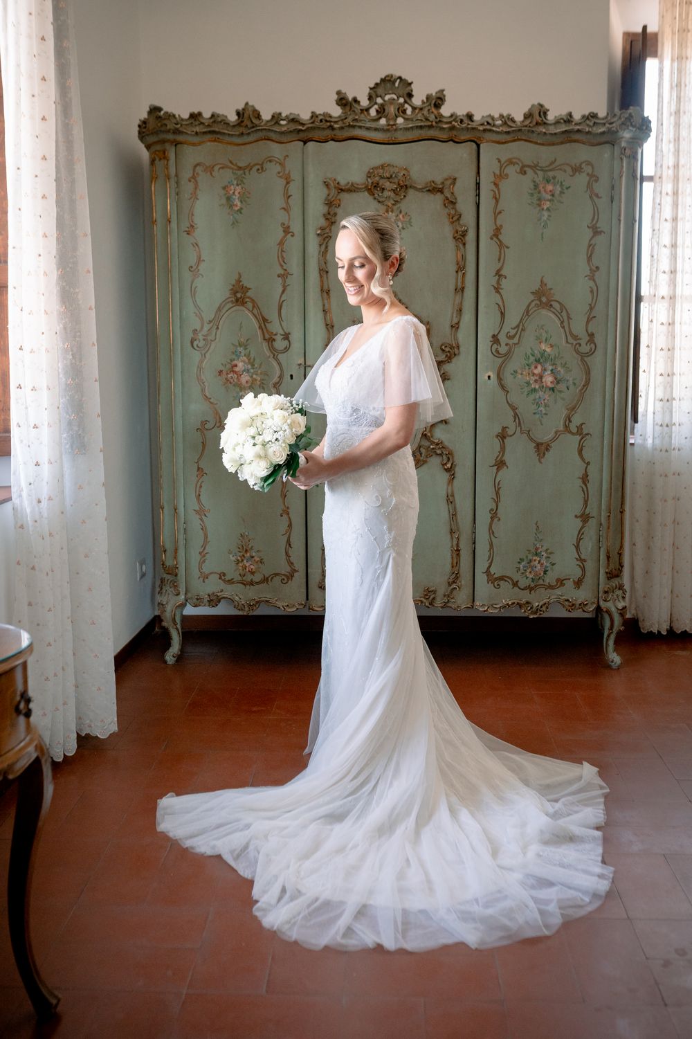 Bride in white gown with train holds bouquet before ornate green antique armoire on terracotta floor.