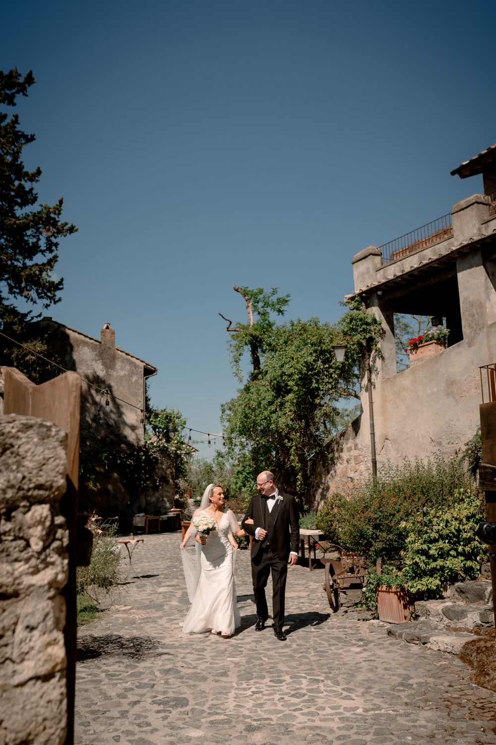 Bride and groom walking hand in hand on cobblestone path surrounded by rustic stone buildings and lush greenery.