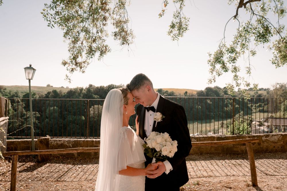 Bride and groom share a romantic moment outdoors, bride holding white bouquet, with scenic countryside views behind them.