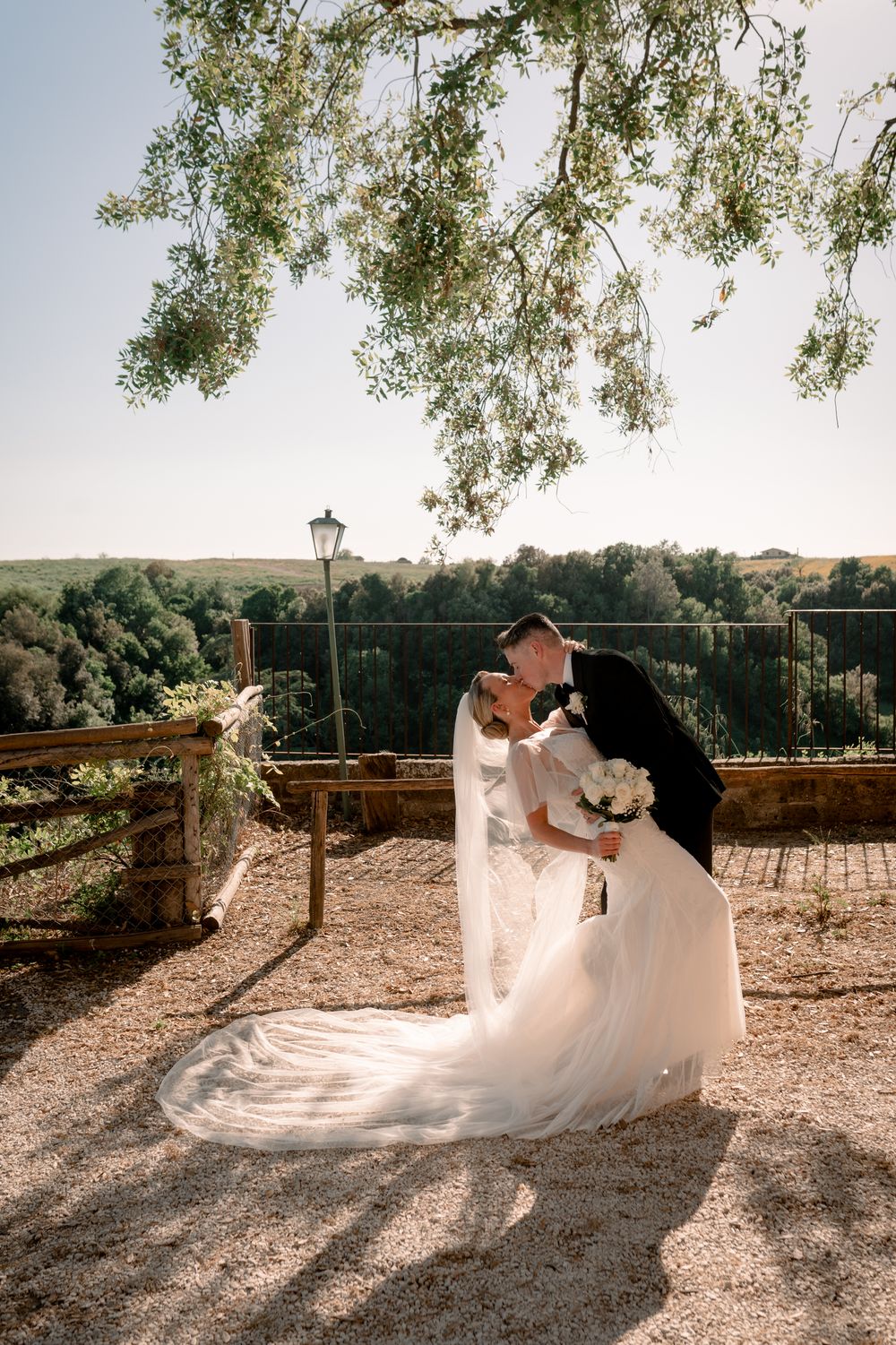 Bride and groom share a romantic kiss outdoors, surrounded by lush greenery, with the bride's long veil flowing gracefully.