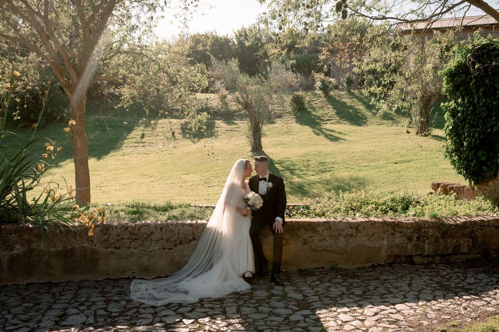 Bride and groom share a romantic kiss at a sunlit stone terrace with lush green gardens in the background.