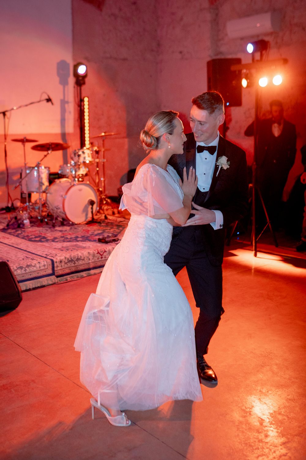 Bride and groom share their first dance at wedding reception with warm red lighting and live band stage in background.