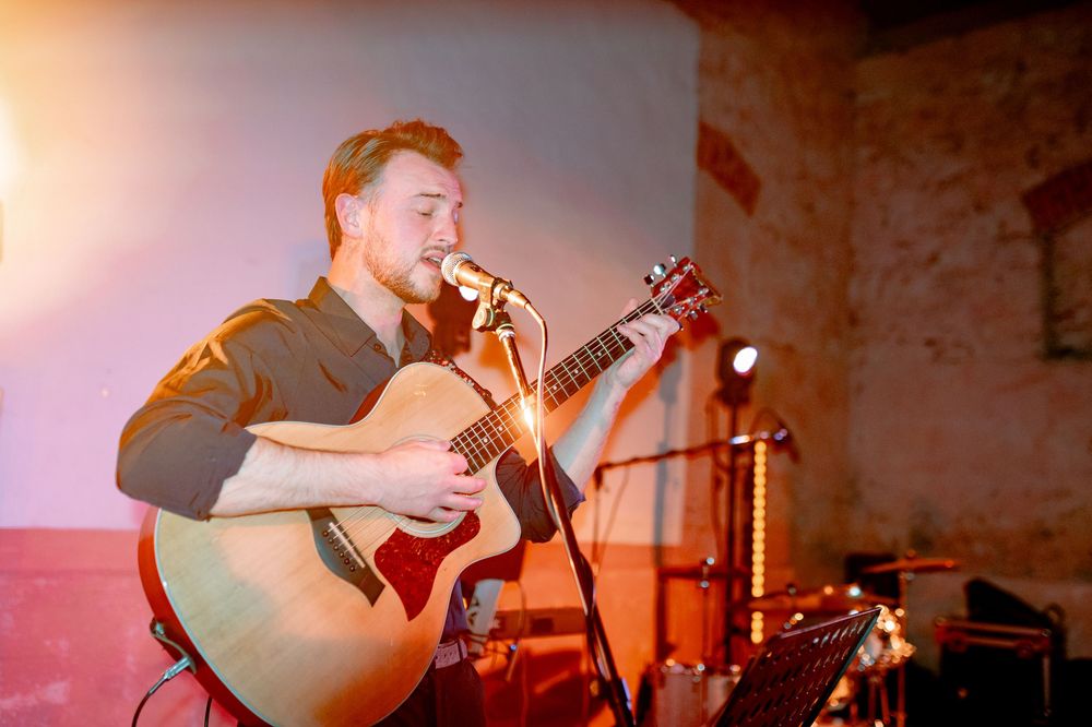 Young male musician playing acoustic guitar and singing into microphone on stage under warm lighting.