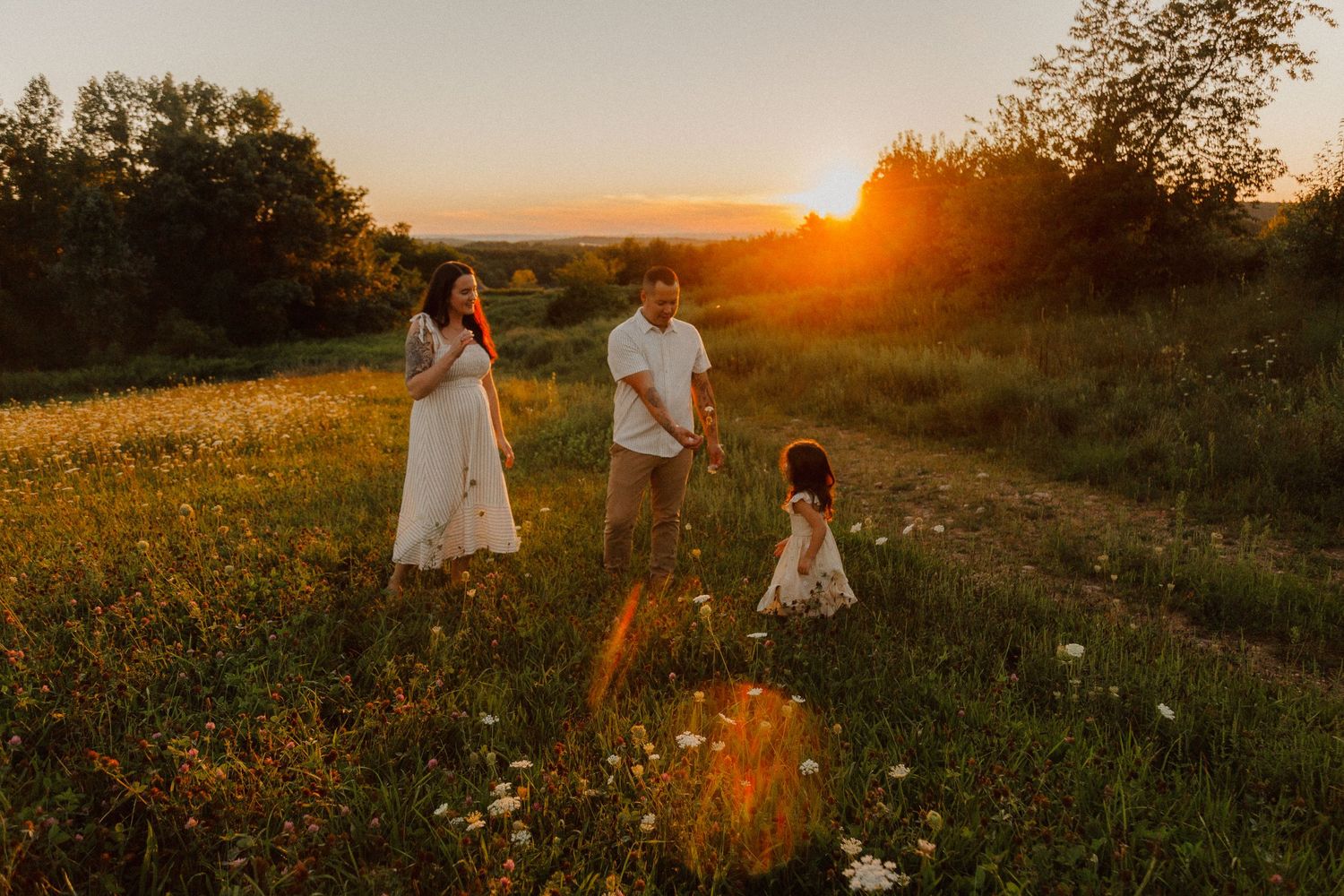Children in white dresses playing in a golden sunlit meadow at sunset with lush green grass and trees.