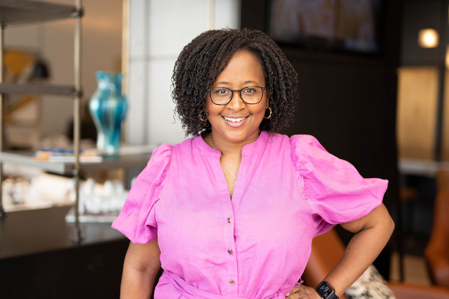 Smiling Black woman in pink blouse with glasses poses confidently in a modern office setting.