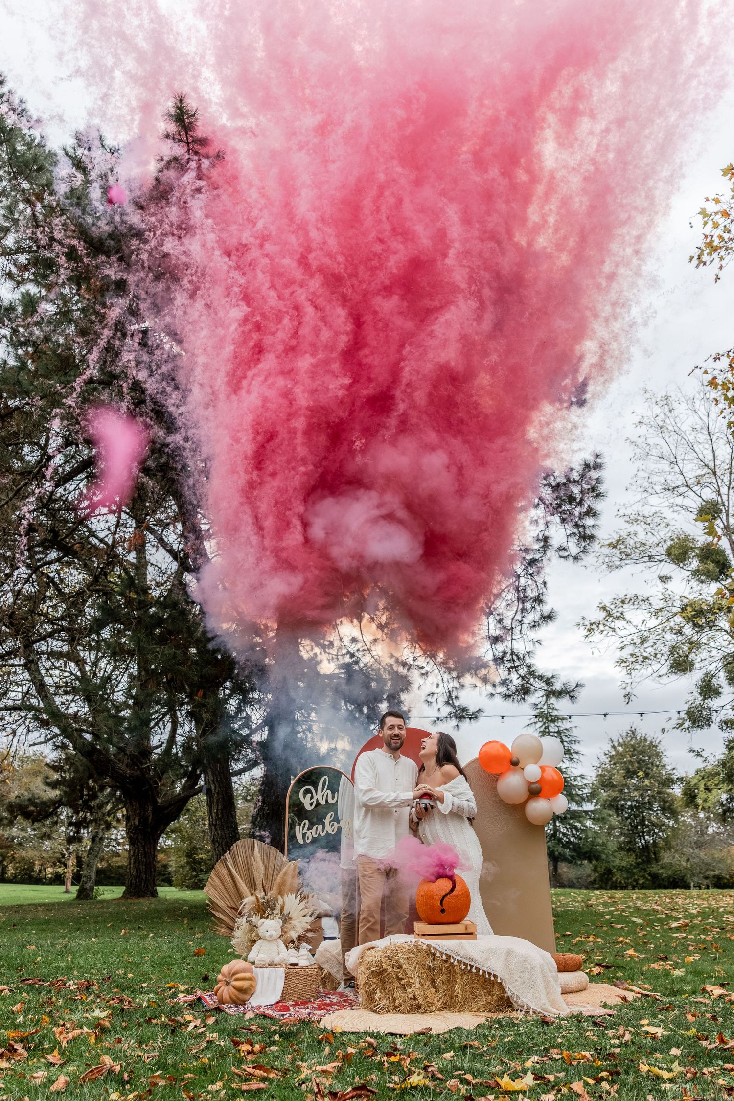 Couple at outdoor gender reveal party with pink smoke bomb, balloon arch, hay bales, and pumpkins in a garden setting.