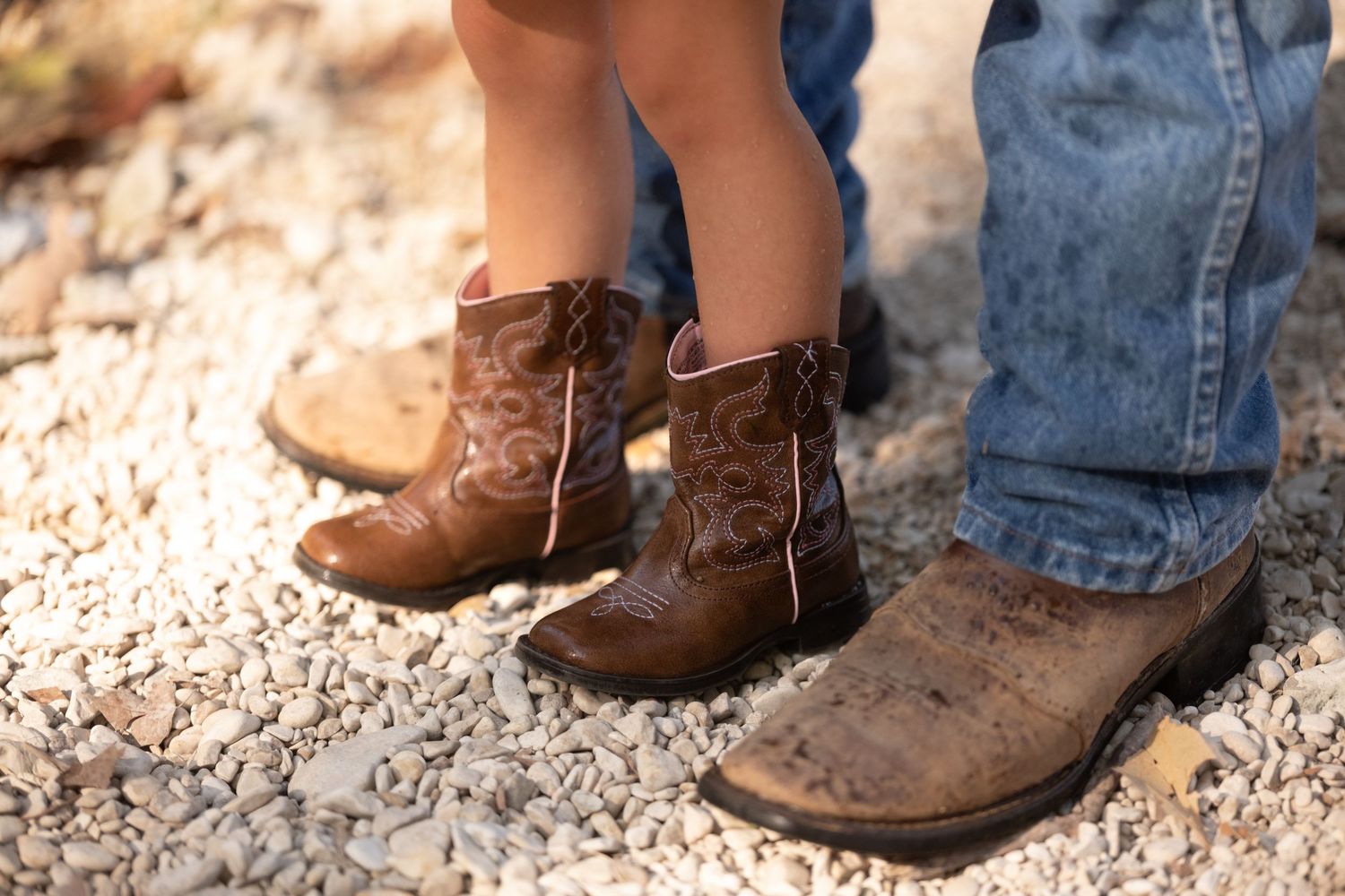 Close-up of adult and child wearing matching brown cowboy boots standing on gravel.
