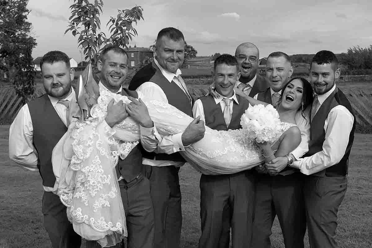 Black and white wedding photo of groomsmen carrying the bride while posing on the lawn of Greenhill Hotel , Wigton, Cumbria