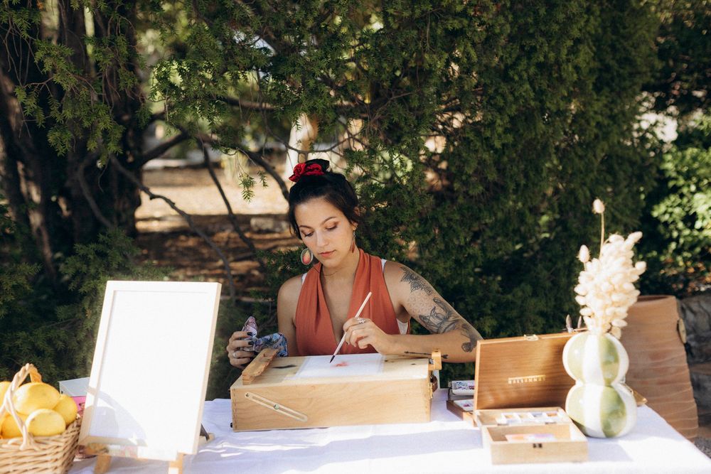 Tattooed woman painting at outdoor table with art supplies, boxes, and vase surrounded by lush green trees.