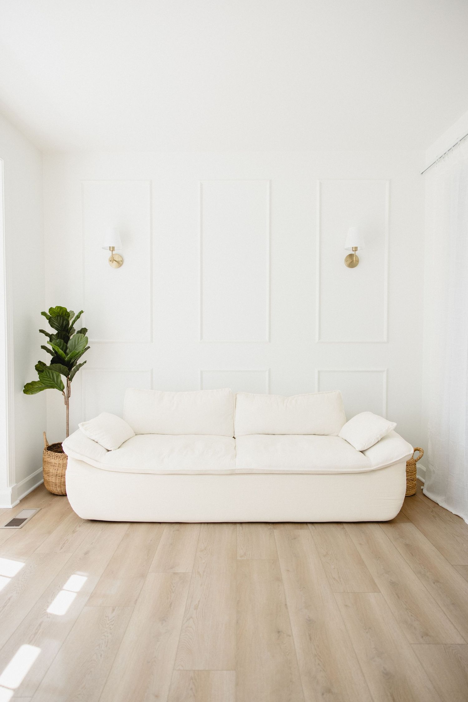 Minimalist white living room with cream sofa, fiddle leaf fig plant, gold wall sconces, and light wood floors.