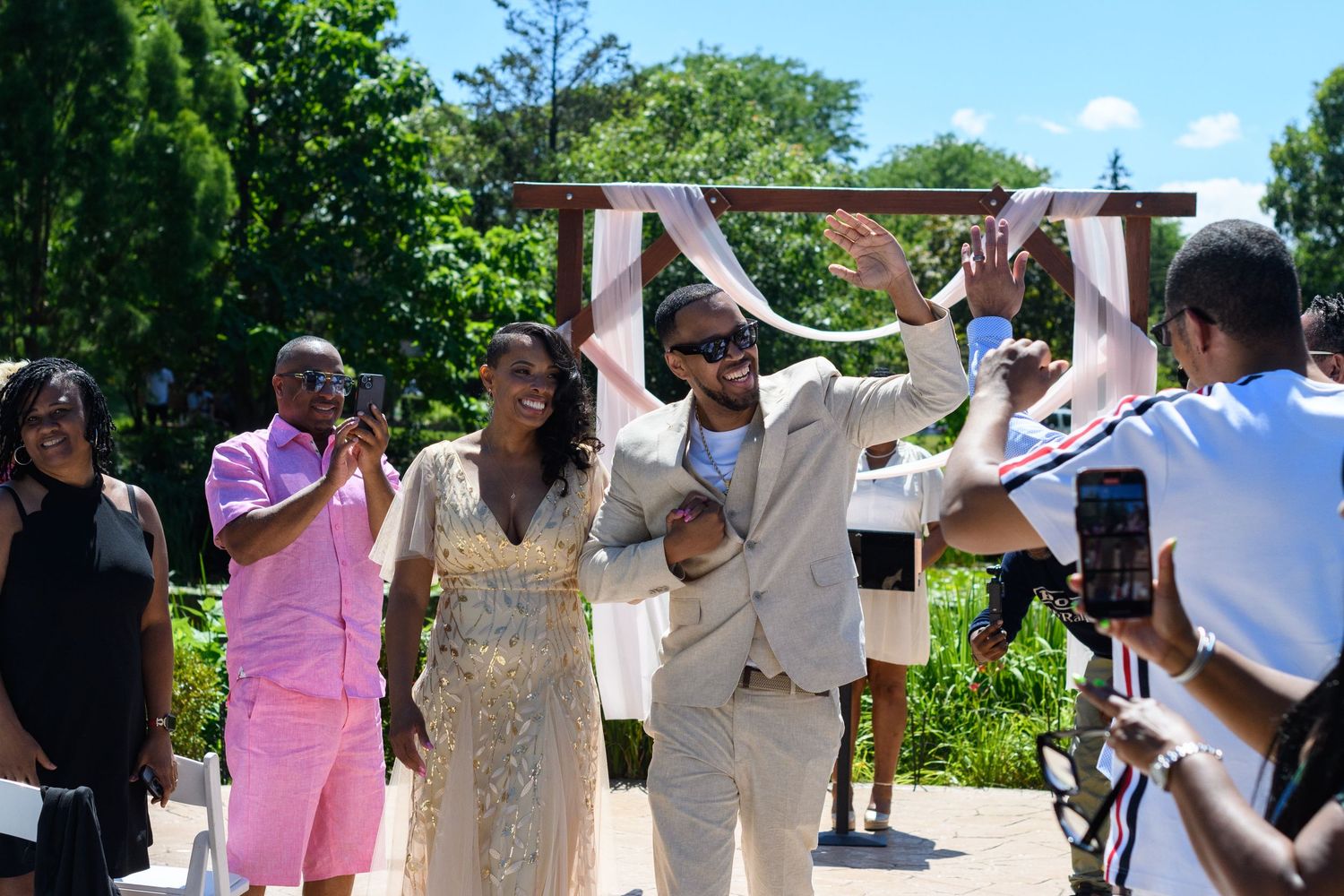 Joyful newlyweds celebrate after outdoor wedding ceremony surrounded by guests in lush green garden setting.