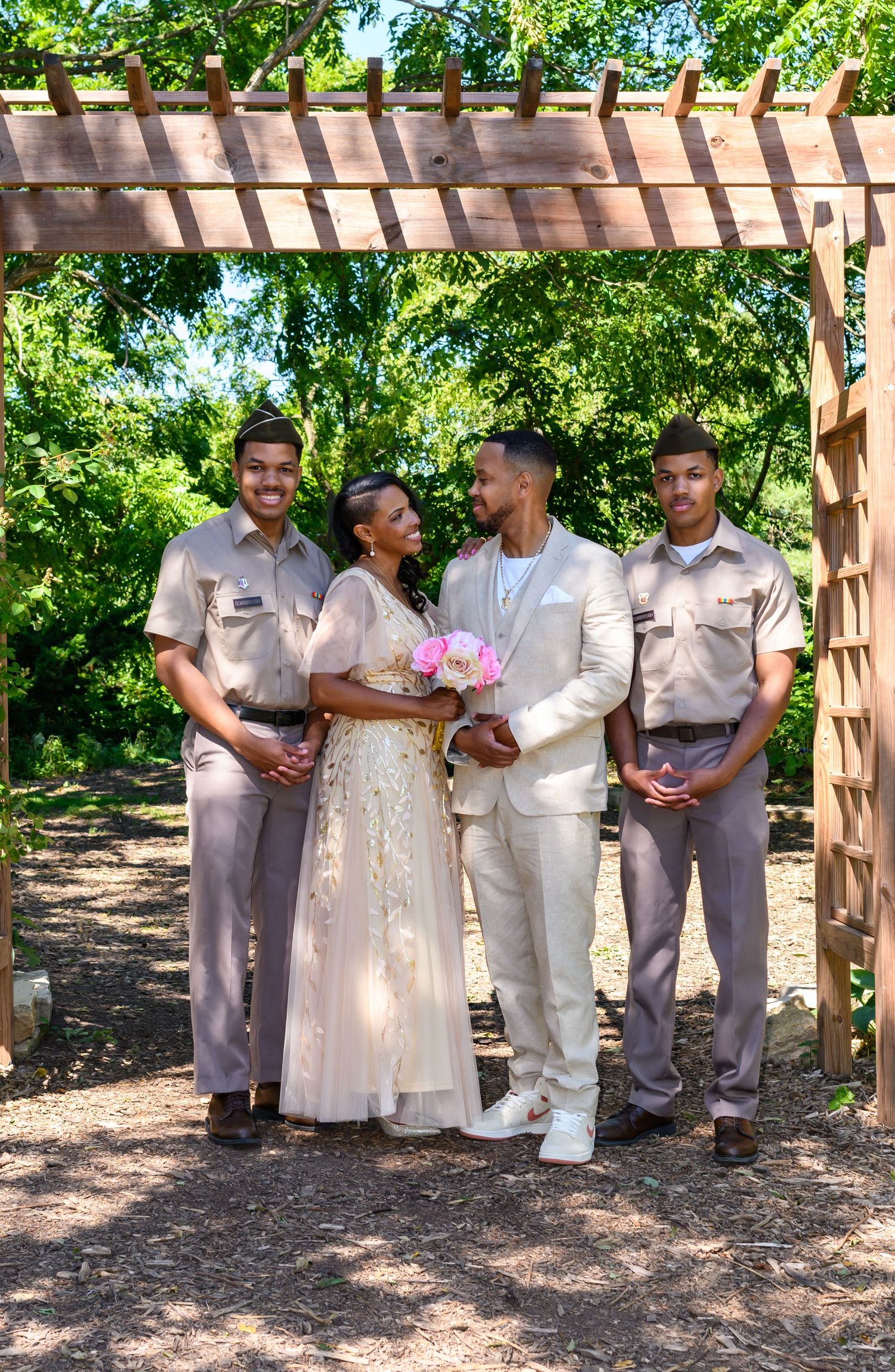 A bride and groom pose with two groomsmen under a wooden pergola surrounded by lush greenery at an outdoor wedding.