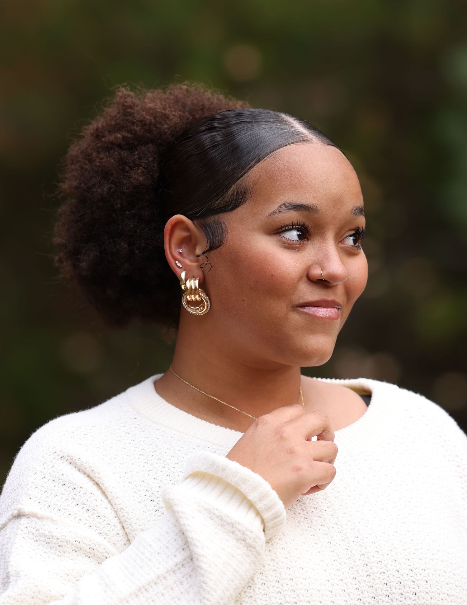 Young woman with curly ponytail wearing gold hoop earrings and white sweater outdoors.