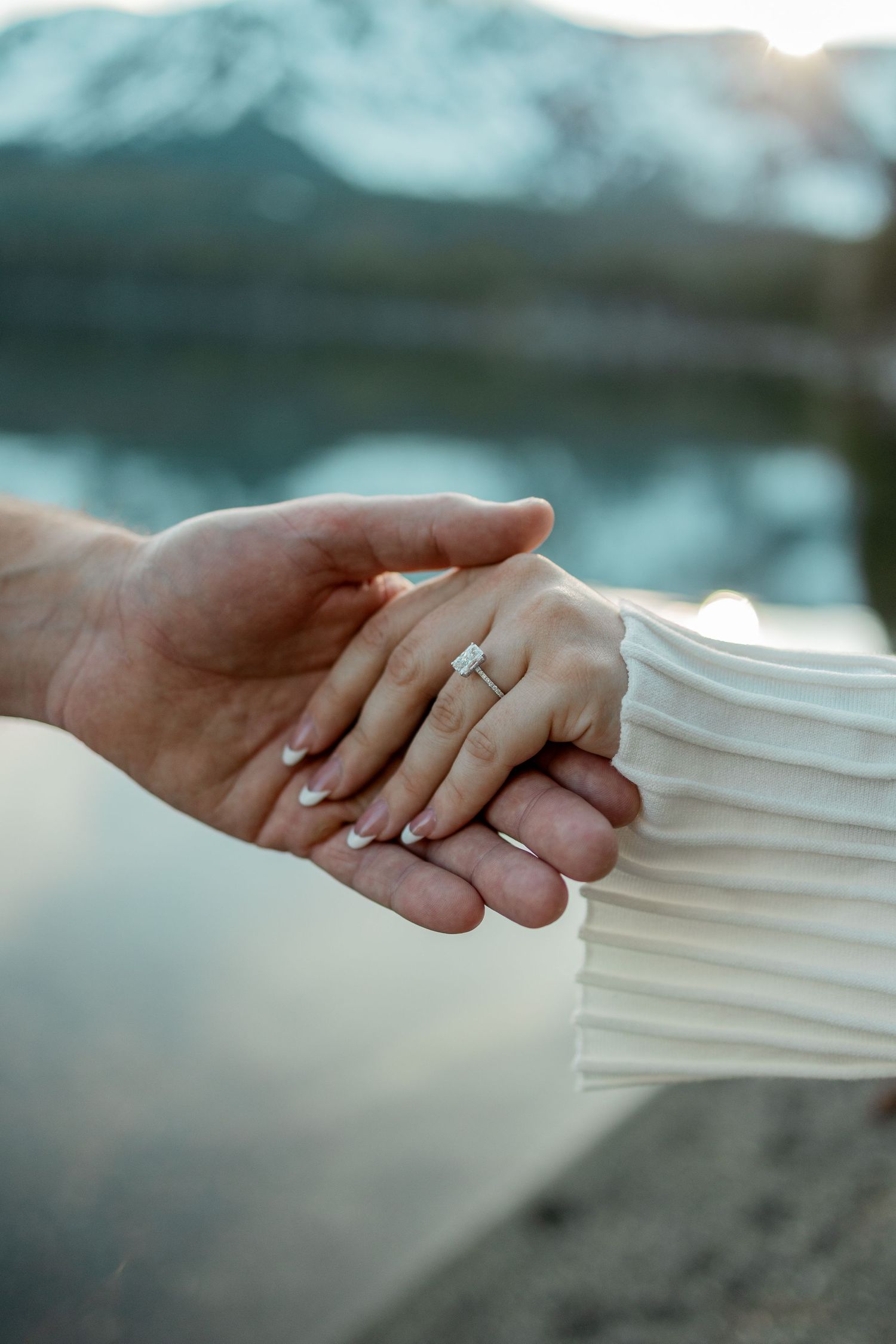 Close-up of couple holding hands with engagement ring, mountain lake backdrop.