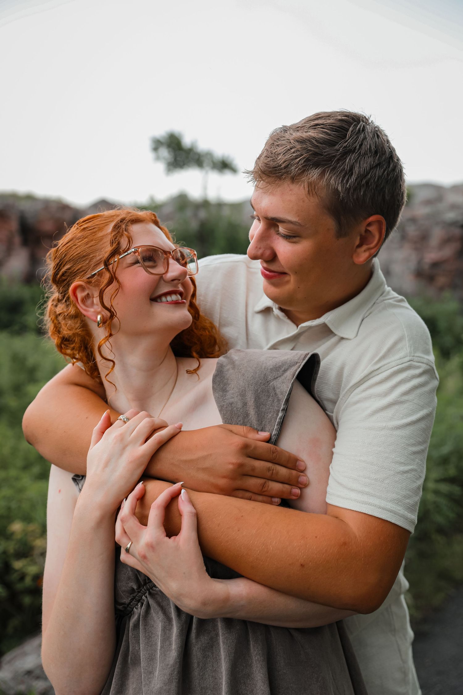 A happy couple embraces outdoors, the redhead woman laughing joyfully as her partner hugs her from behind.