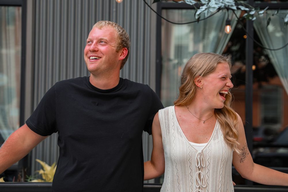 A smiling couple laughing together outdoors, man in black tee and woman in white lace top.