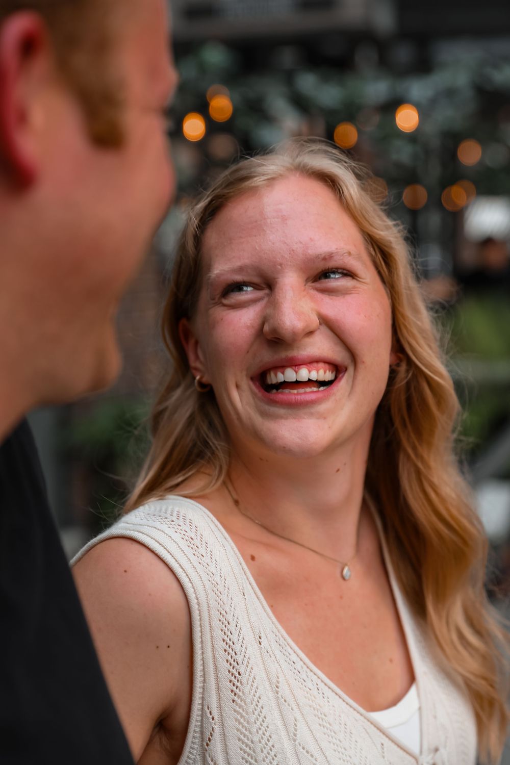 A young woman with long blonde hair laughs joyfully outdoors, wearing a white knit vest with bokeh lights behind her.