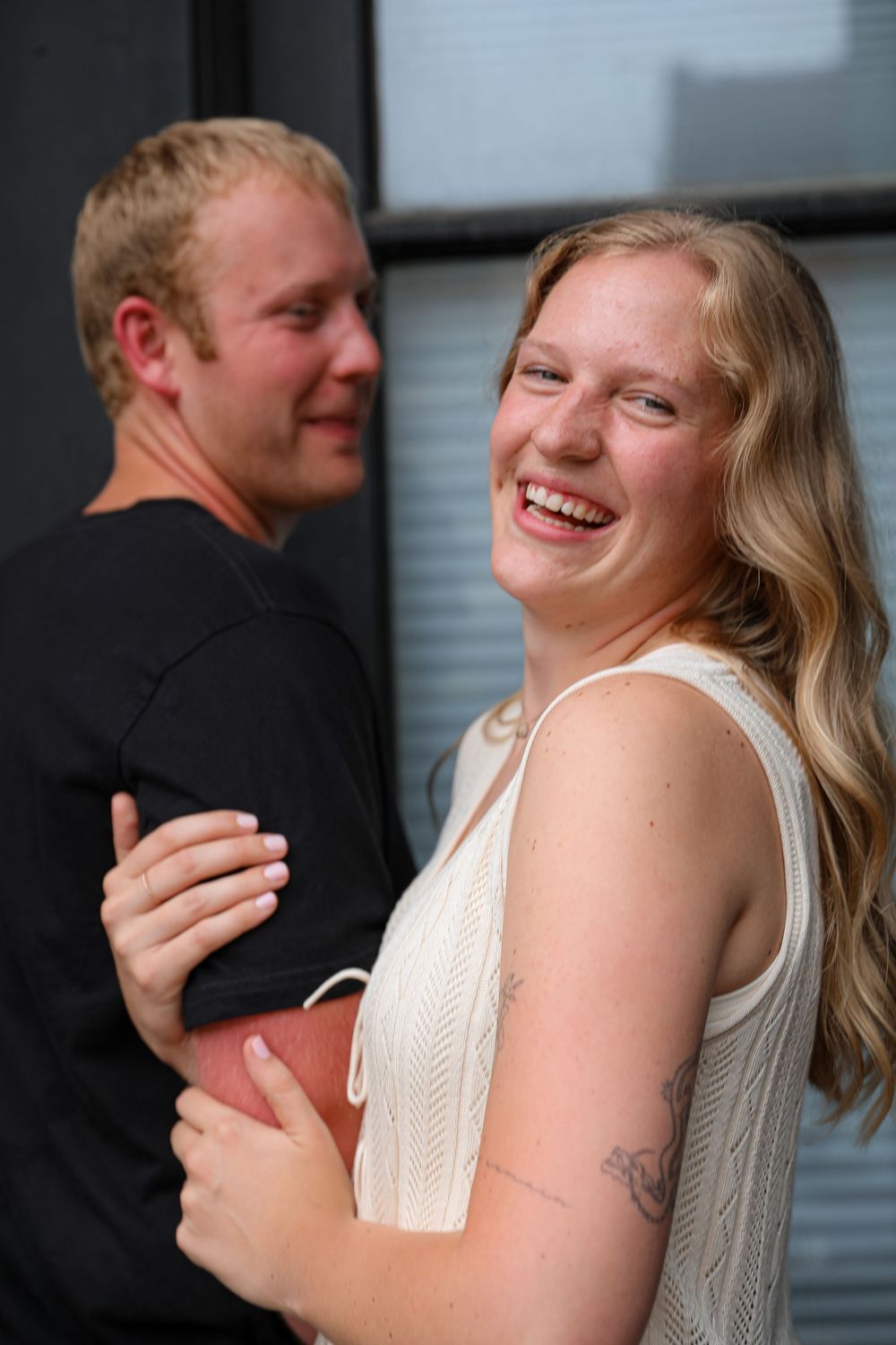 Smiling blonde woman with tattoo embraces man in black shirt outdoors.
