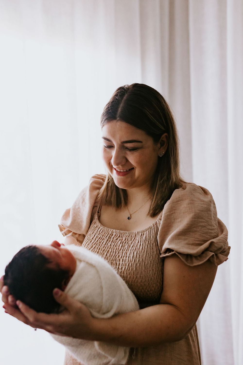 Smiling young mother tenderly holding her newborn baby wrapped in white swaddle near a bright window.