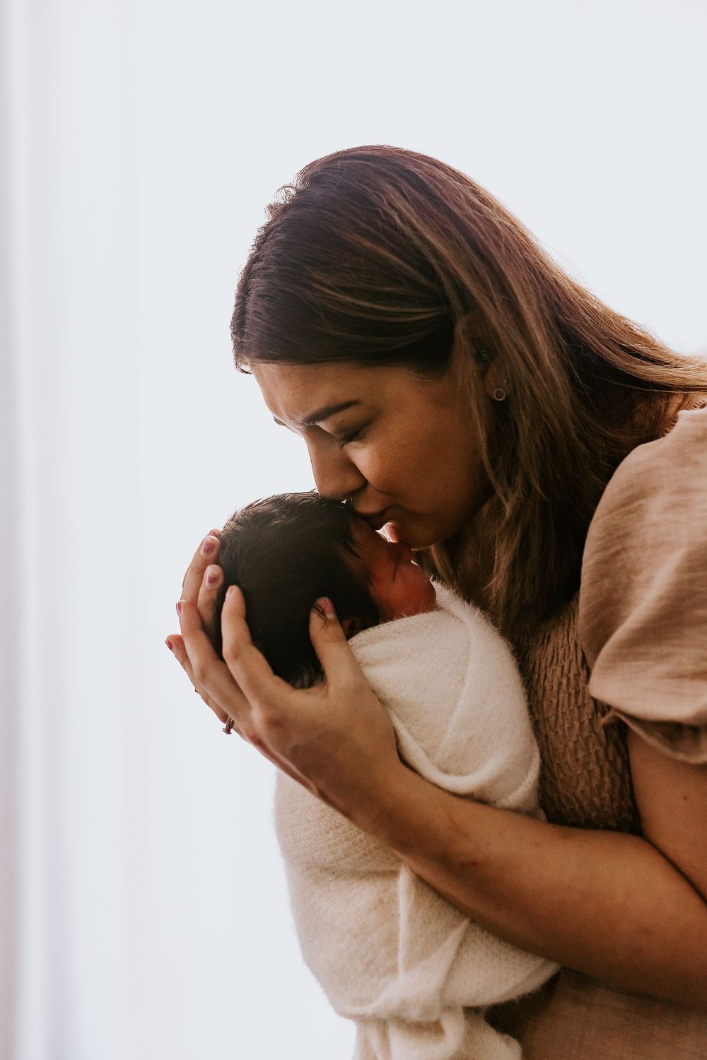 A mother tenderly kisses her newborn baby wrapped in a white blanket near a bright window.