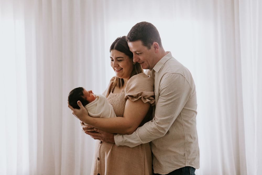 A couple smiles lovingly at their newborn baby wrapped in a white swaddle during a soft, natural light newborn photo session.