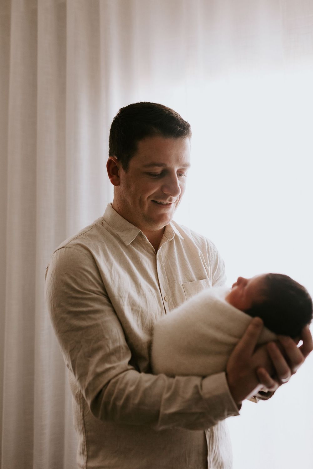 A smiling father tenderly holds his swaddled newborn baby against a soft white curtain backdrop.