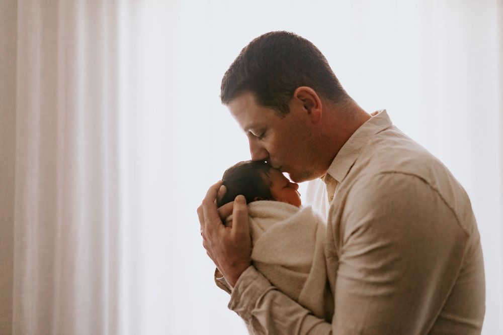 Loving father kissing his newborn baby wrapped in a cozy blanket near a bright window.