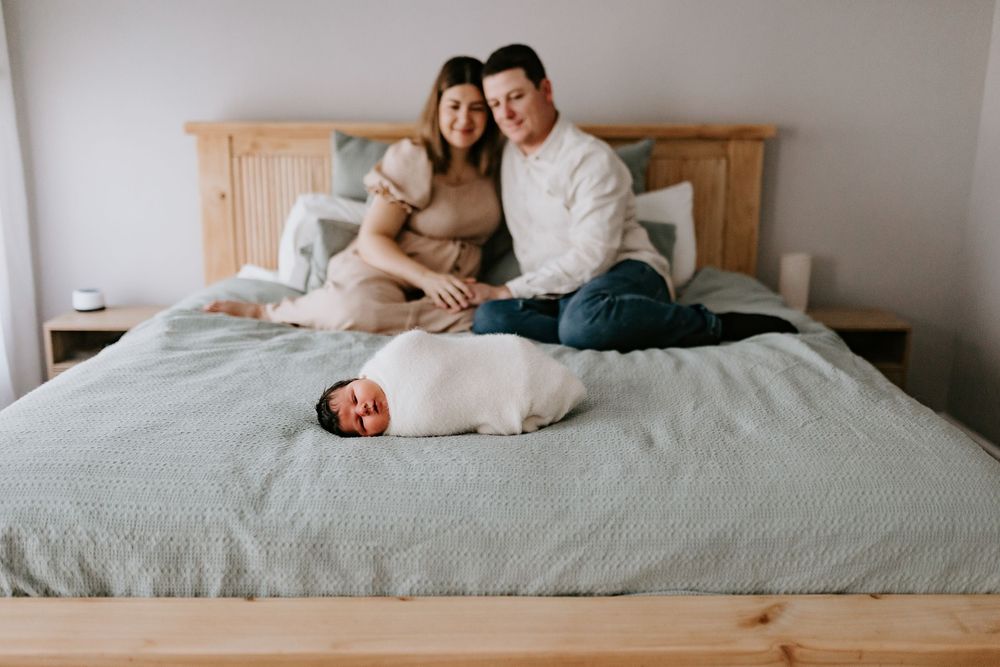 A couple sits on a bed smiling at their swaddled newborn baby lying in front of them.