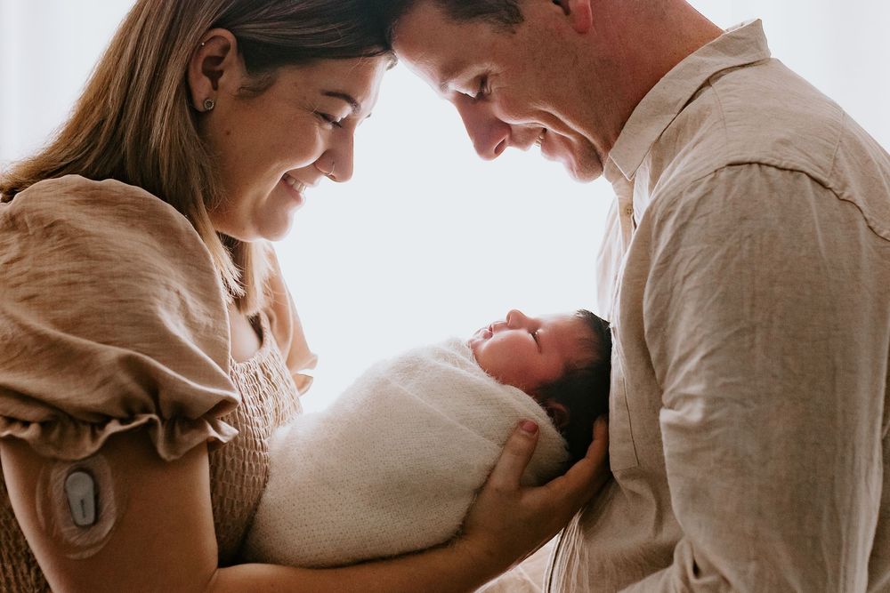 Happy parents smiling at their newborn baby wrapped in white swaddle in a warm, softly lit portrait.