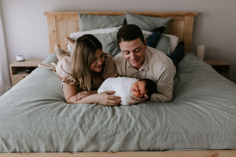 New parents lying on bed smiling at their swaddled newborn baby during an in-home lifestyle photo session.