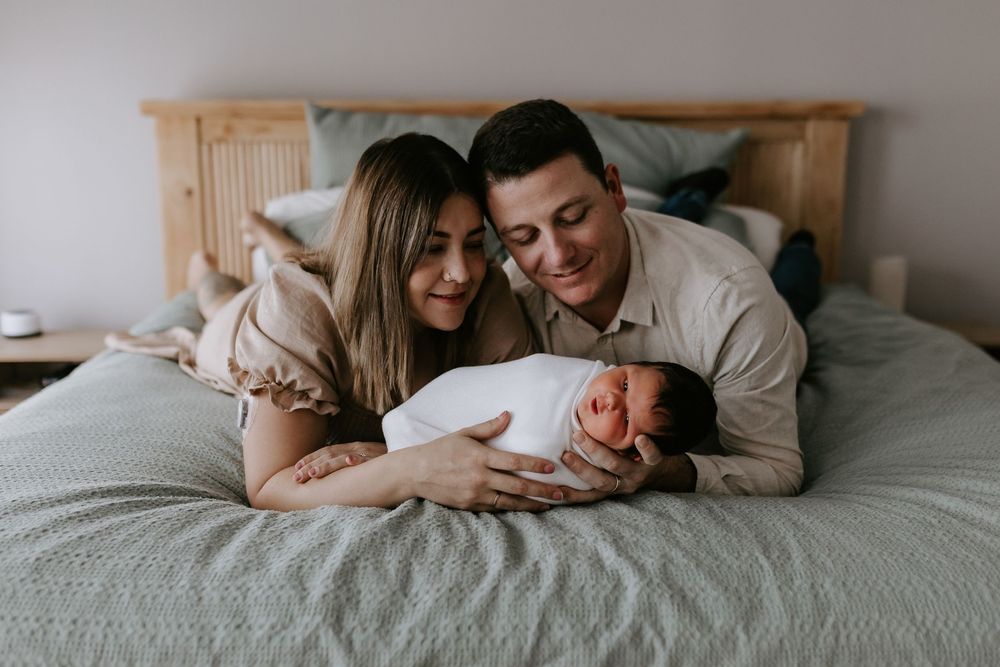 A couple lying on a bed smiling at their newborn baby wrapped in white, capturing a tender family moment.
