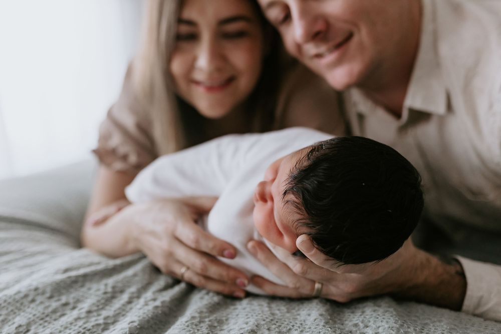 Parents smiling lovingly at their newborn baby wrapped in white, lying on a soft gray blanket.