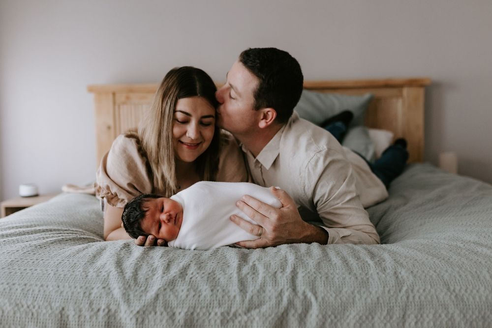Happy couple lying on bed smiling at their newborn baby wrapped in white swaddle blanket.