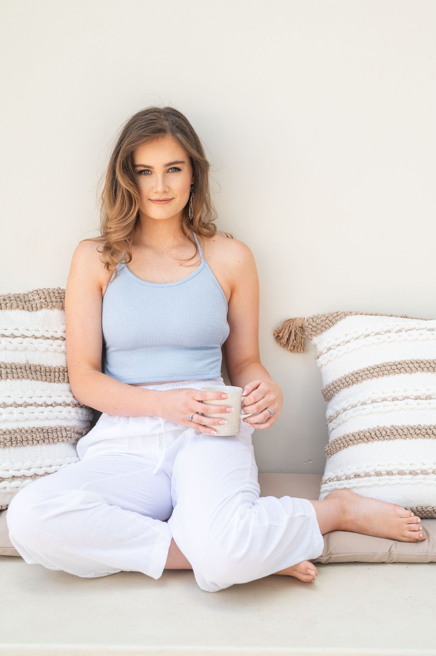 Woman in light blue tank top and white pants relaxing on cushions against a white wall.