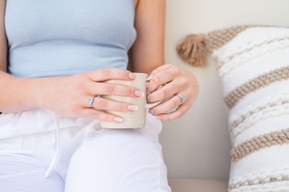 Woman in white pants holding a small ceramic mug with manicured nails and rings, sitting on a couch.