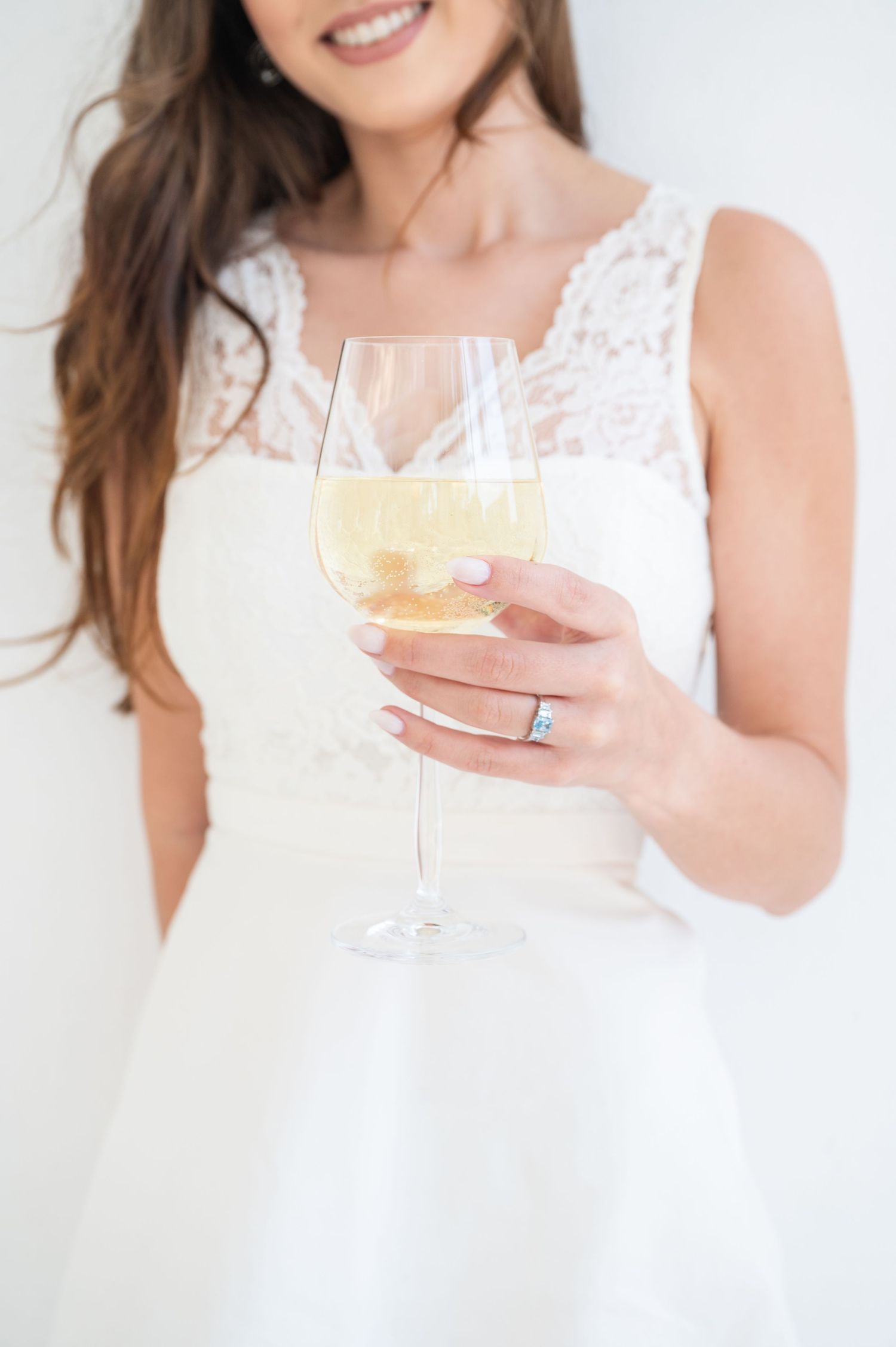 Person in white lace dress holding a champagne glass with bubbling golden liquid against light background.