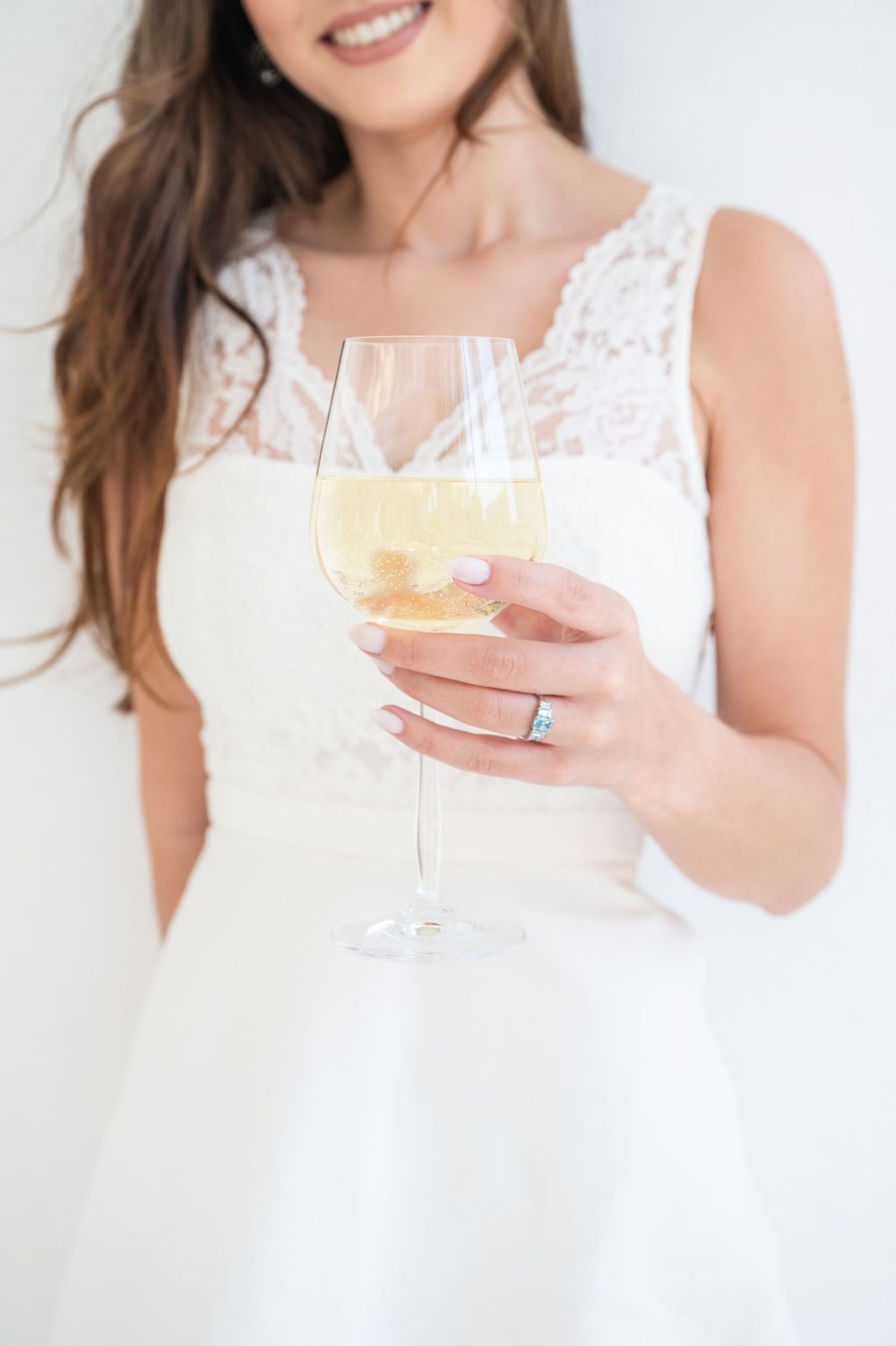 Person in white lace dress holding a champagne glass with bubbling golden liquid against light background.