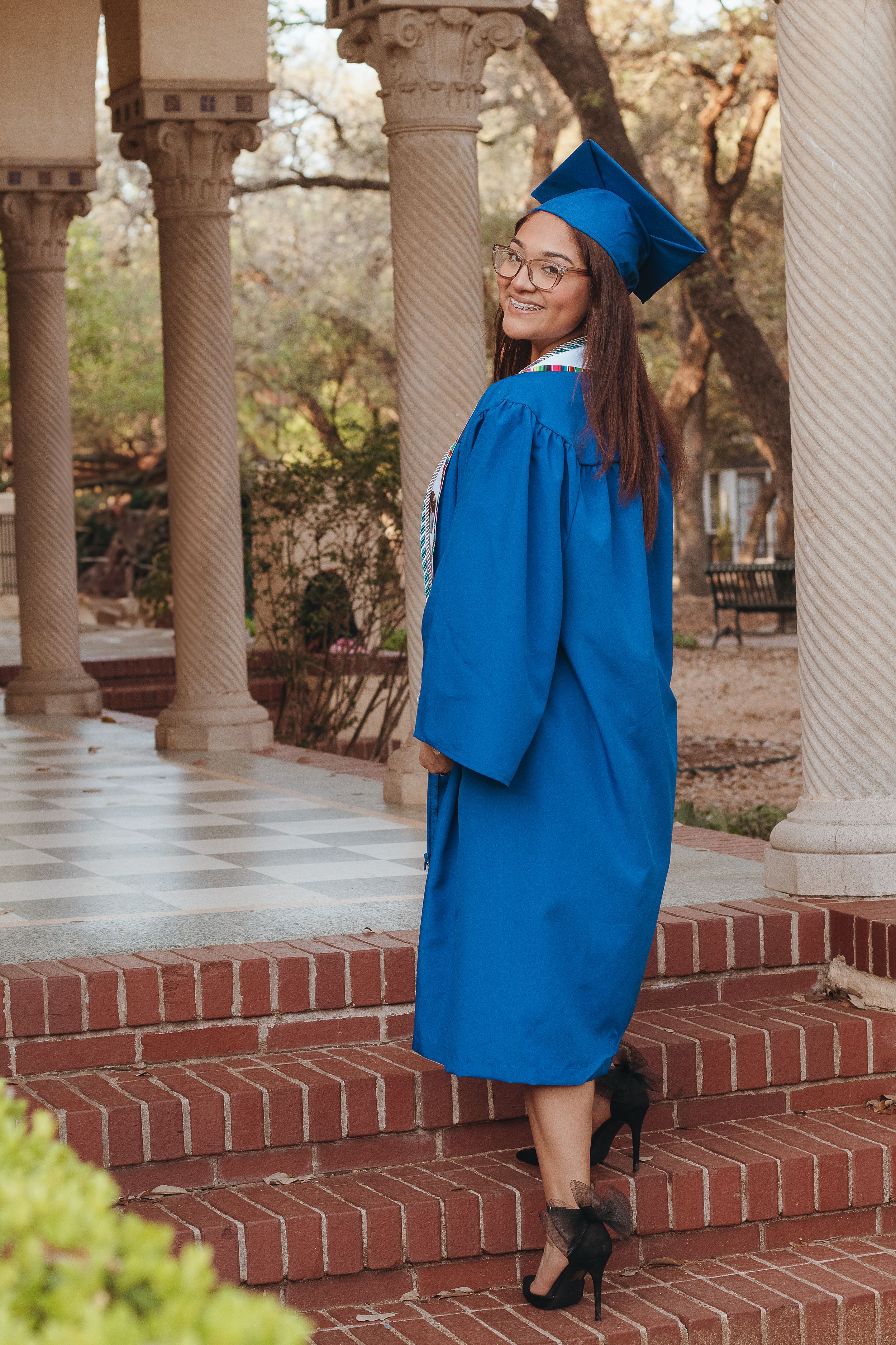 Young woman in blue graduation cap and gown smiling over shoulder on brick steps between stone columns.