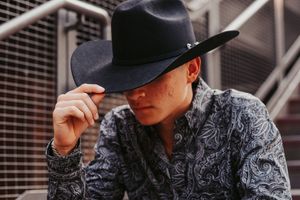 Young man in a black cowboy hat and paisley shirt tilting his brim outdoors.