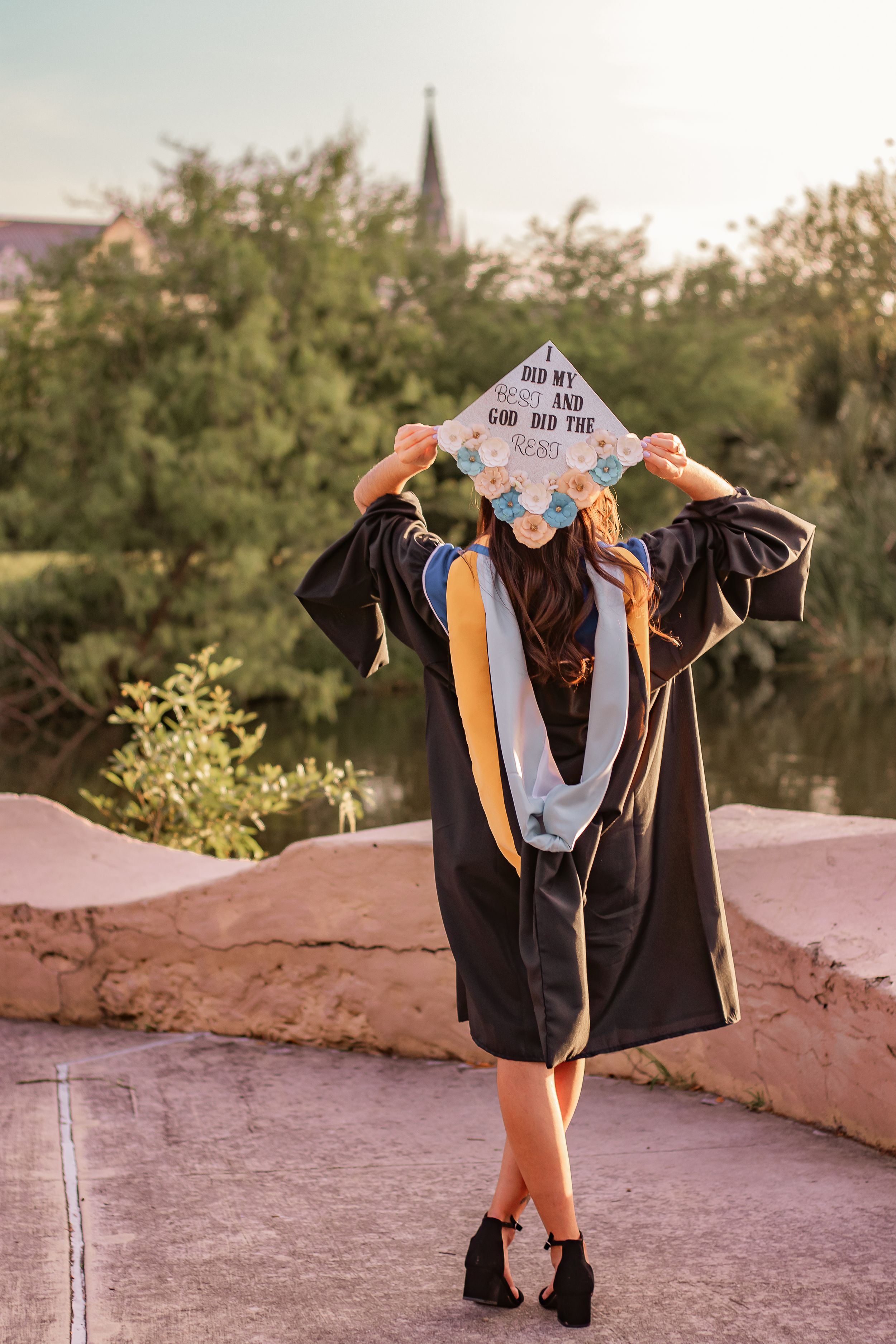 Graduate in black cap and gown holds decorated mortarboard reading inspirational quote outdoors near water.