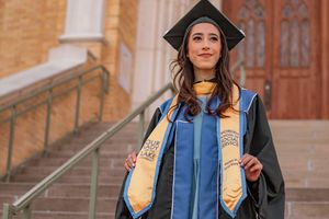 Young woman in graduation cap and gown smiling on university steps during commencement ceremony.