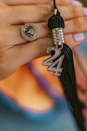 Hand holding a graduation tassel with a 2024 charm and wearing a class ring with lavender nail polish.