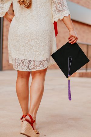 Woman in white lace dress holding black graduation cap with purple tassel, wearing red wedge sandals.