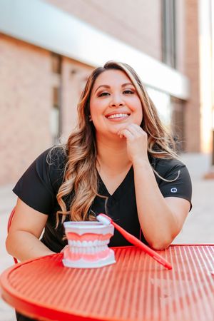 Smiling dental professional in black scrubs sitting outdoors at red table with dental model and toothbrush.