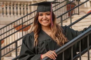 Smiling young woman in black graduation cap and gown posing on steps at her college commencement ceremony.