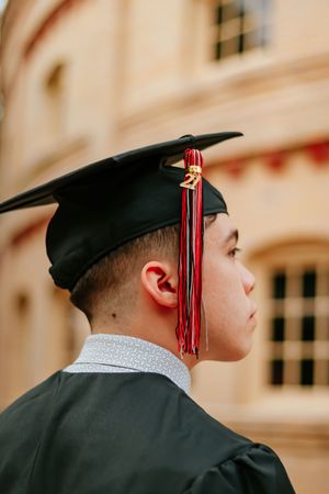 Young male graduate in black cap and gown with red tassel, viewed from behind, standing outdoors.