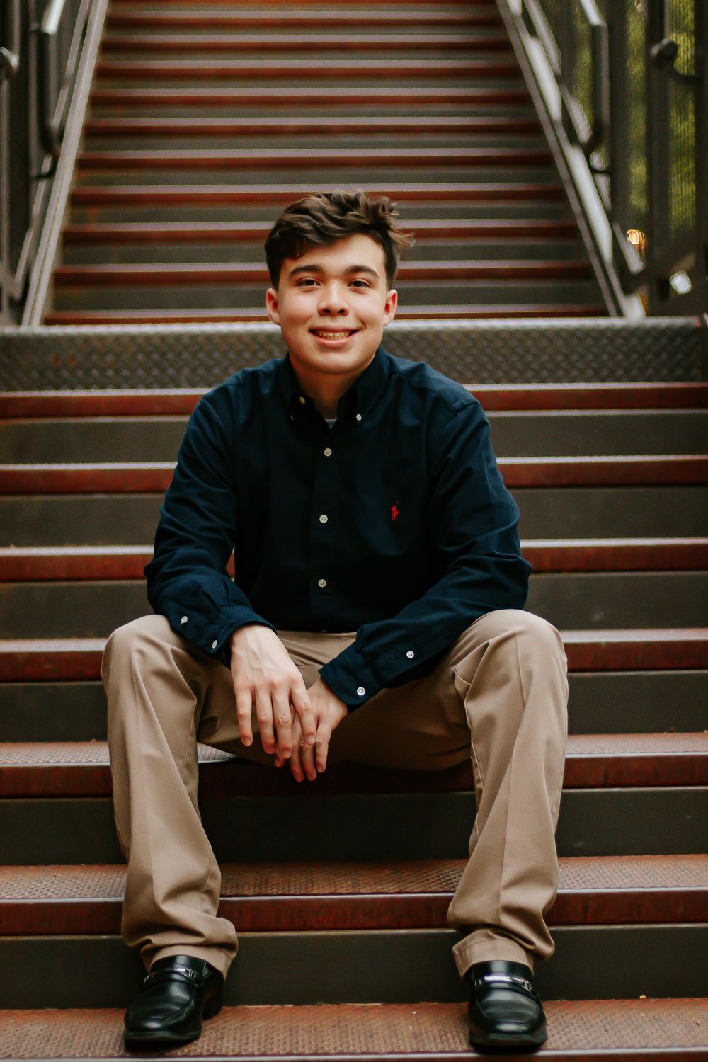 Young man in navy Polo shirt and khakis sitting on outdoor stairs, smiling confidently at camera.